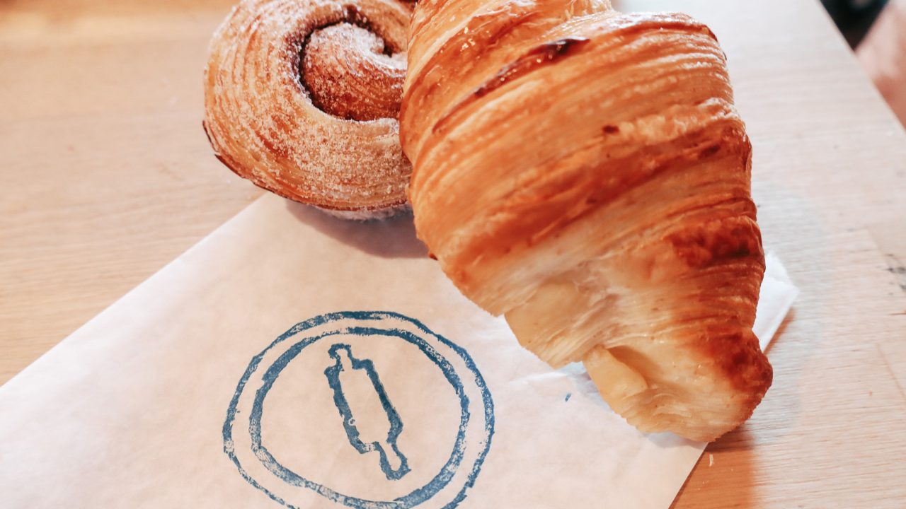 A croissant and a spiral pastry rest on a white paper with a blue circular logo, placed on a light wooden surface.