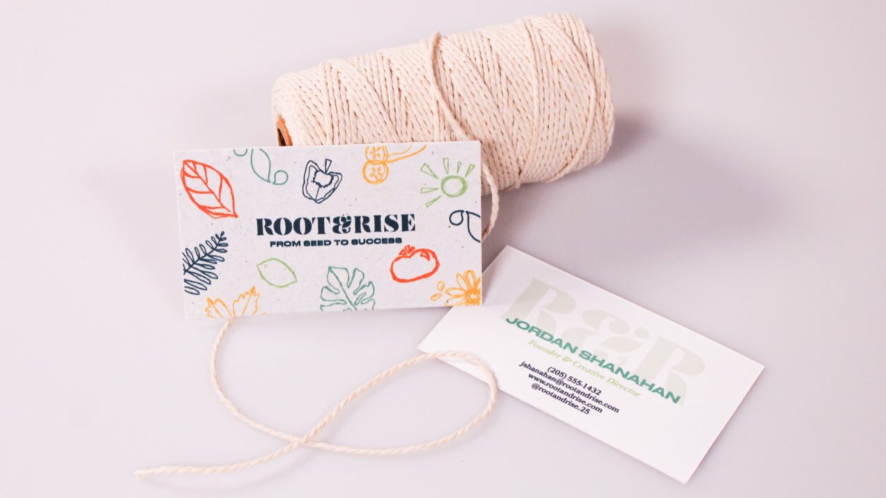 A spool of white string with two tags attached; one tag has colorful illustrations and says ROOT & RISE, the other is a business card for Jordan Shanahan.