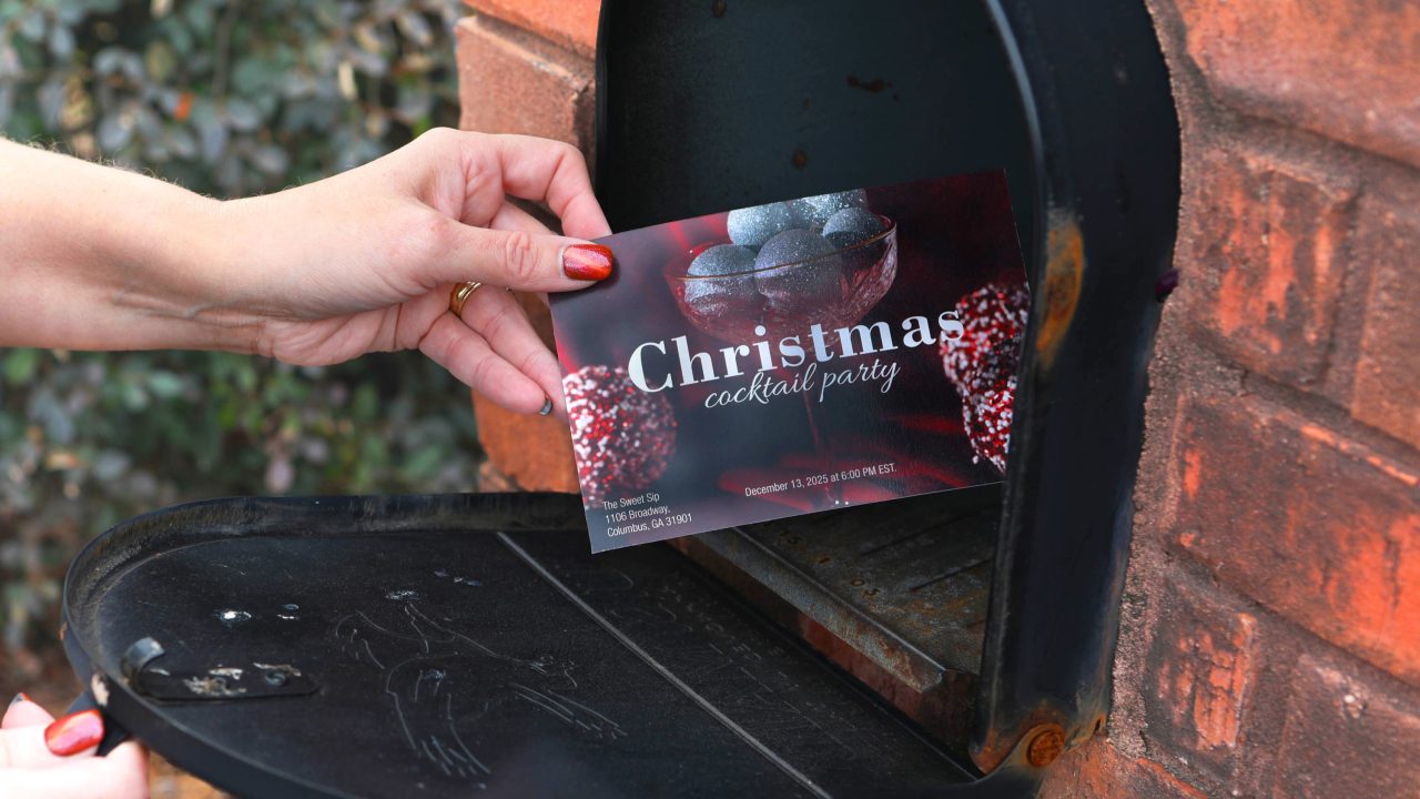 A hand places a Christmas cocktail party invitation into a black mailbox attached to a brick wall.