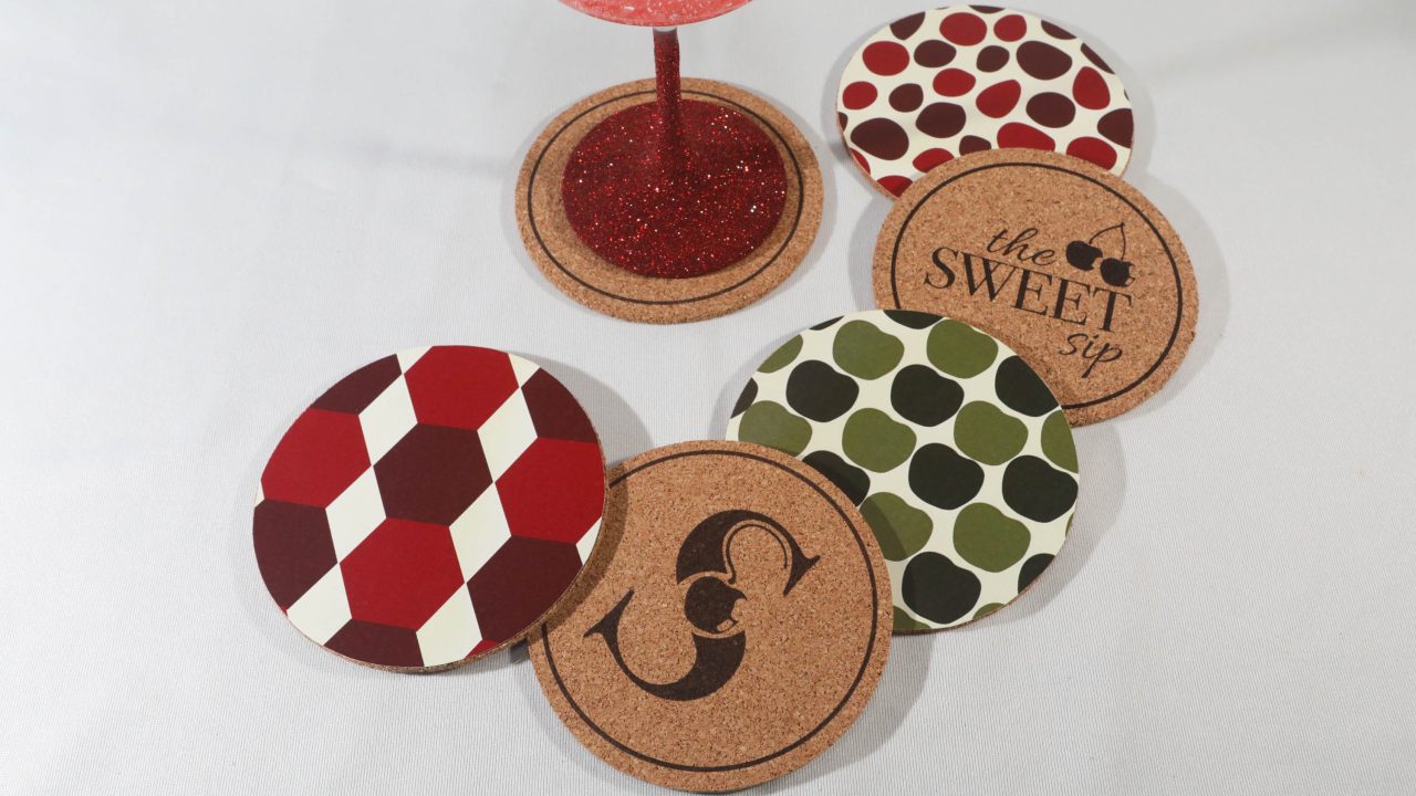 A red glitter wine glass on a cork coaster, with four other cork coasters nearby, each featuring different geometric or apple-themed patterns.