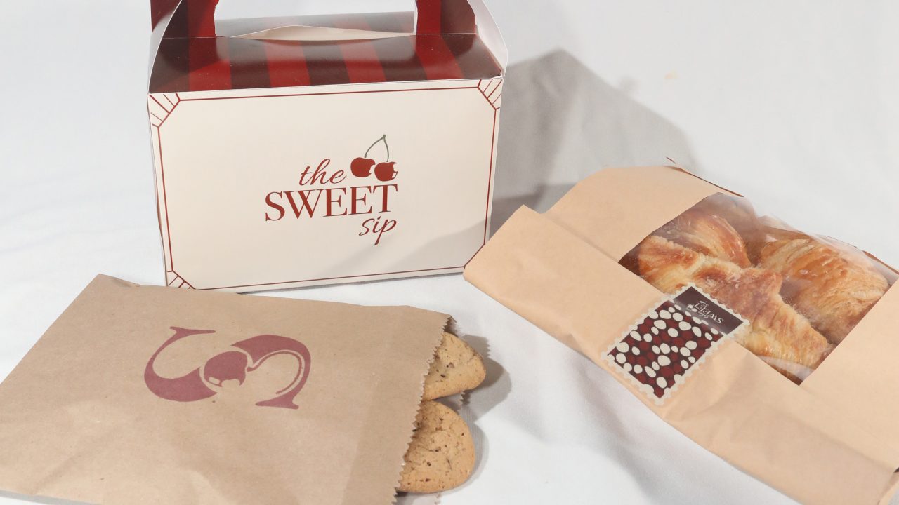 A box labeled The Sweet Sip, a paper bag with two cookies, and a paper sleeve holding several croissants are displayed on a white surface.