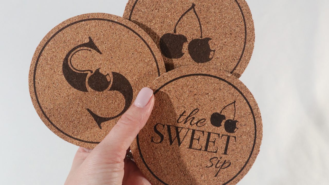 A hand holds three round cork coasters, each with a black cherry-themed design and the words the SWEET sip printed on one of them.