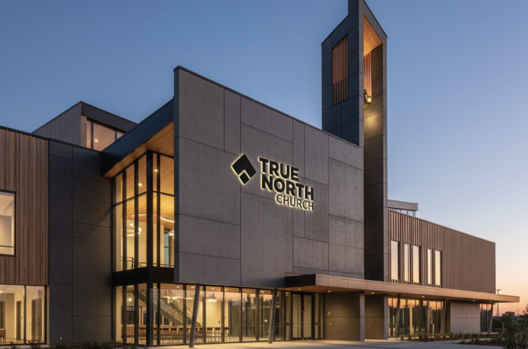 Modern church building with large windows and a sign reading True North Church illuminated at dusk.