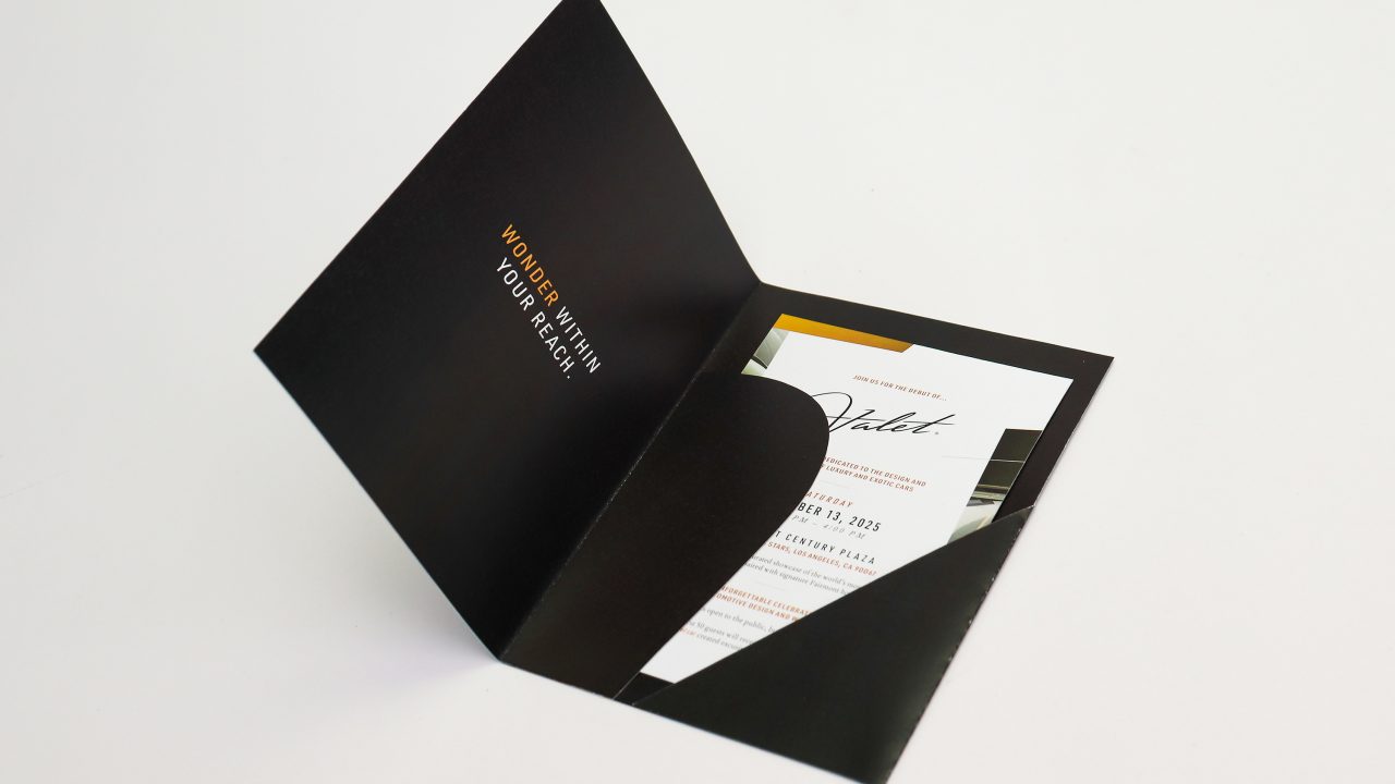 A black folder partially open, revealing an invitation card inside with printed text and a bold Valet script in the center.