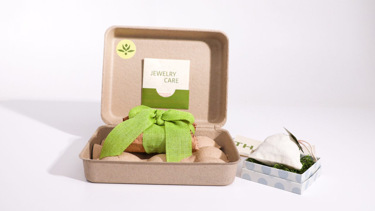 A small jewelry box with a green ribbon, an eco-friendly care card, and a white heart-shaped pouch placed next to it on a white surface.