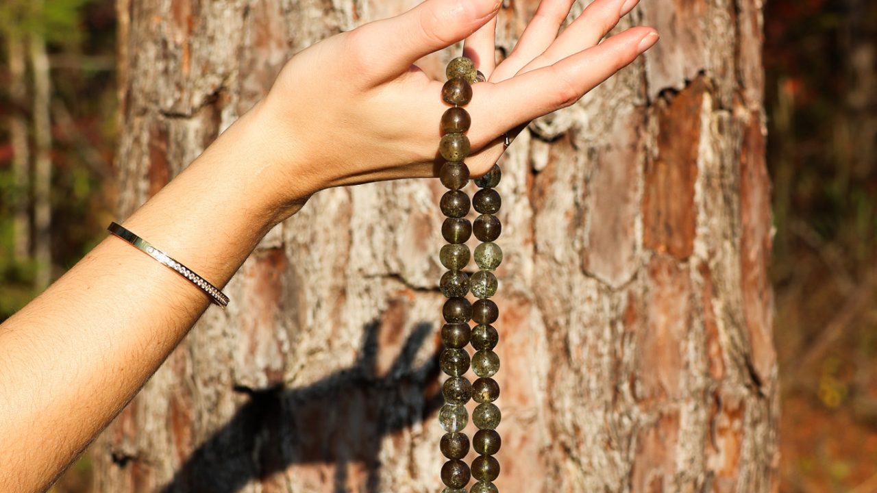 A hand holding a beaded green necklace is positioned in front of a tree trunk. Sunlight casts a shadow of the necklace onto the tree bark.