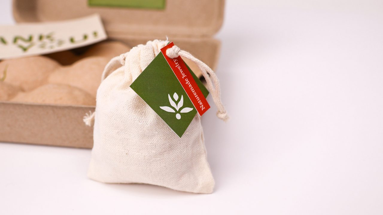 A small drawstring cloth bag with a green tag featuring a leaf design sits in front of a cardboard box on a white background.
