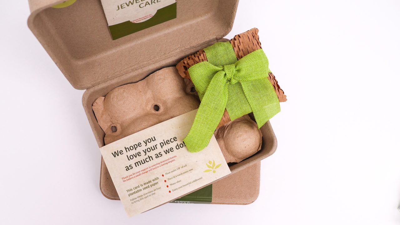 An open eco-friendly box contains a card, a small woven package tied with a green ribbon, and protective packaging materials.