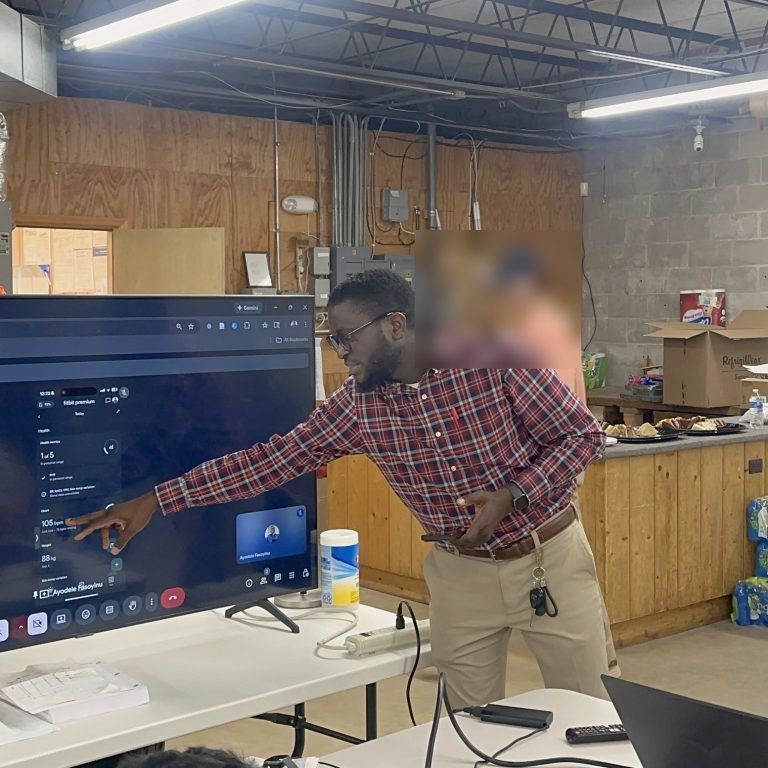 A man in a plaid shirt points at a large screen during a presentation in an industrial-style room with supplies and boxes in the background.