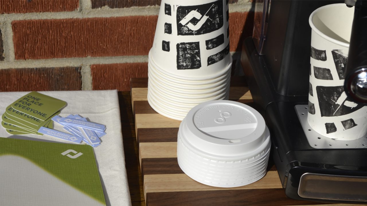 A stack of paper cups, plastic lids, and sugar packets sits on a wooden tray next to a coffee machine and a stack of green note cards.