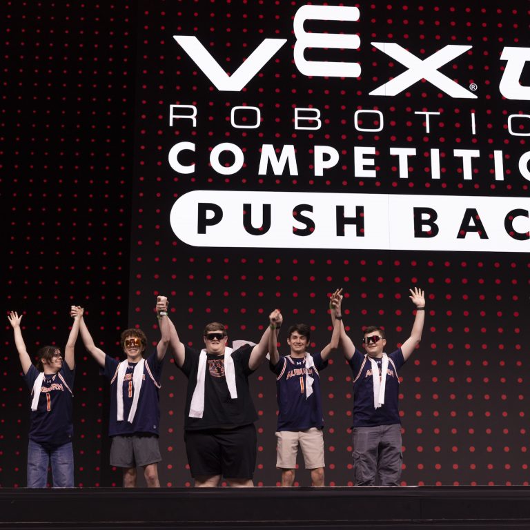 Five people wearing medals stand on stage, raising their hands, in front of a large VEX Robotics Competition Push Back sign with a red dotted background.