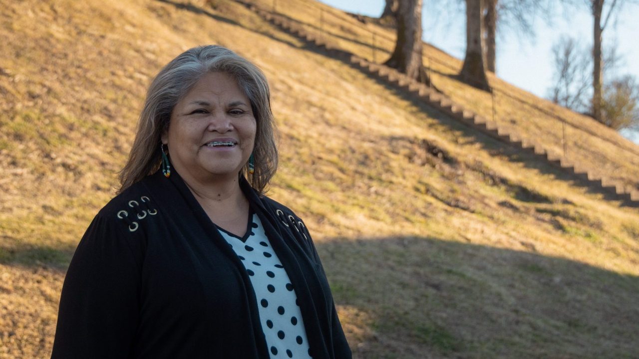 A woman with gray hair stands outdoors in front of a grassy hill with bare trees and a long staircase.