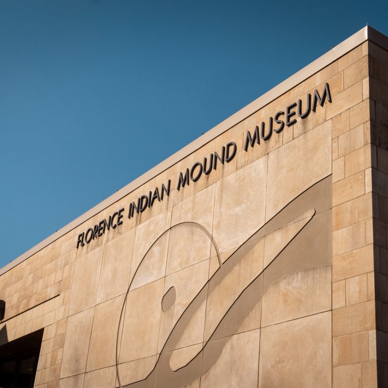 Exterior view of the Florence Indian Mound Museum building, featuring its name and a large abstract mural on the tan stone facade under a clear blue sky.