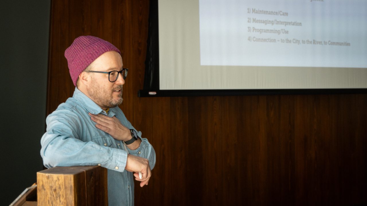 A person in a red beanie and glasses stands at a podium, gesturing, with a presentation slide projected on a screen behind them.