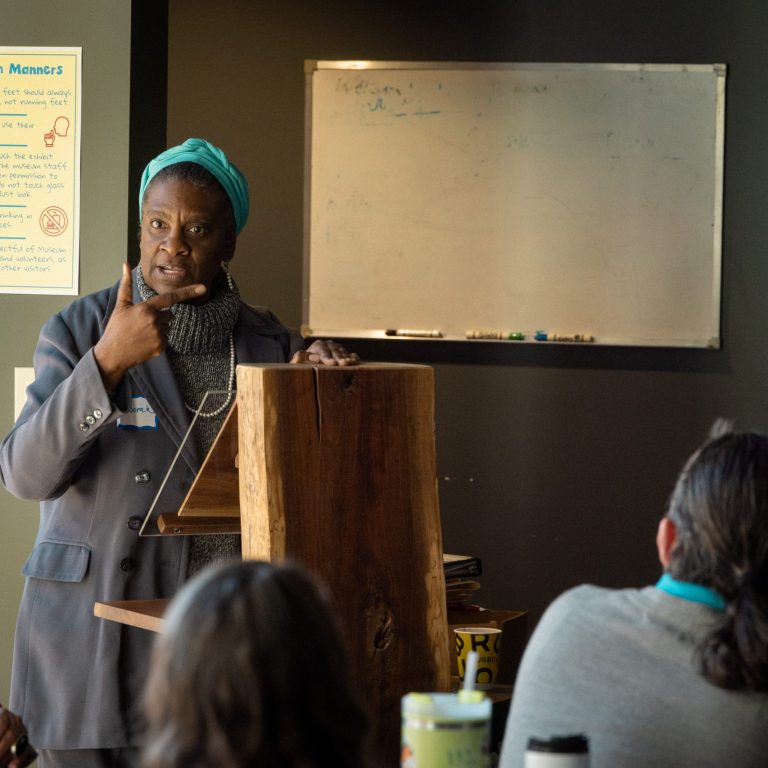 A woman stands at a wooden podium speaking to an audience in a classroom, gesturing with her hand. A whiteboard and a poster are visible in the background.