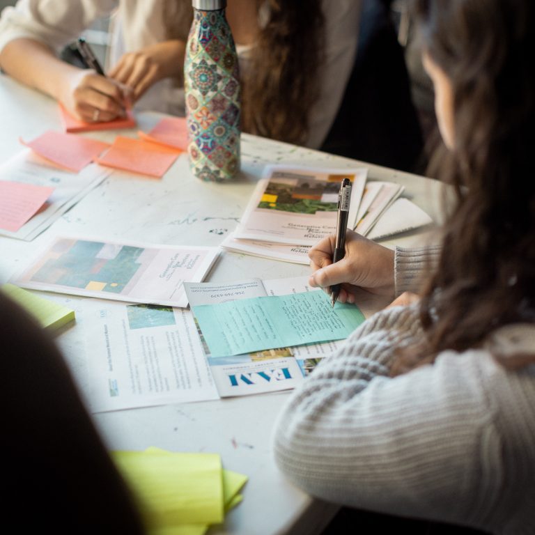 People sitting at a table writing on colored paper and sticky notes, with brochures, a patterned water bottle, and scattered documents visible.