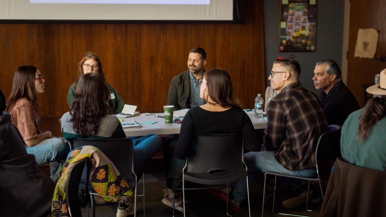 A group of people sit in a circle having a discussion in a room with a projector screen and wooden walls.
