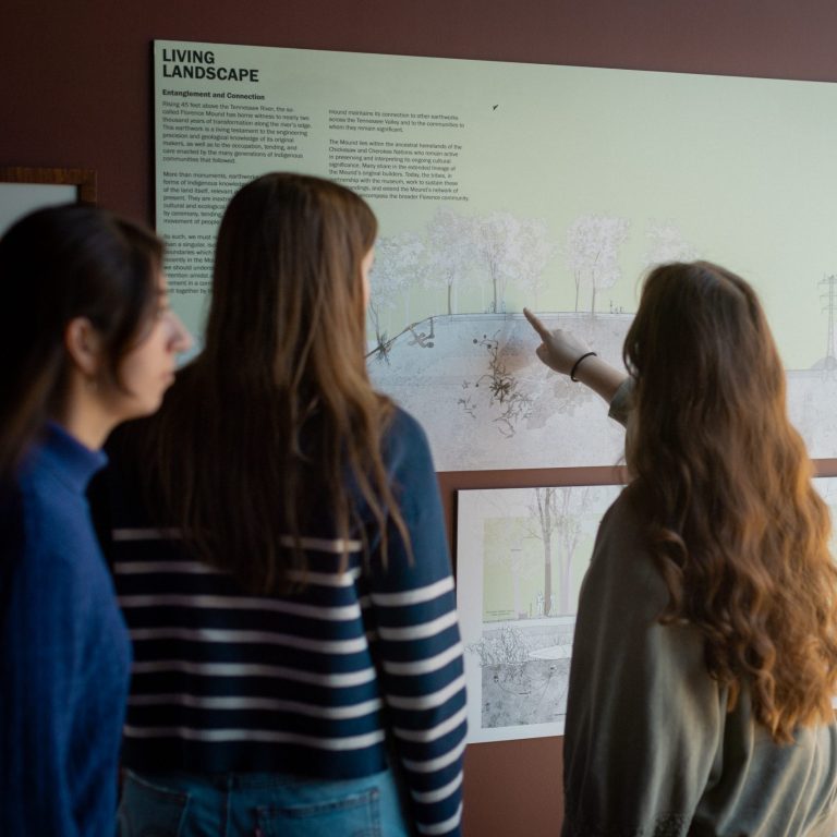 Three people stand indoors, looking at and pointing toward an informational display titled Living Landscape with diagrams and text on a wall.