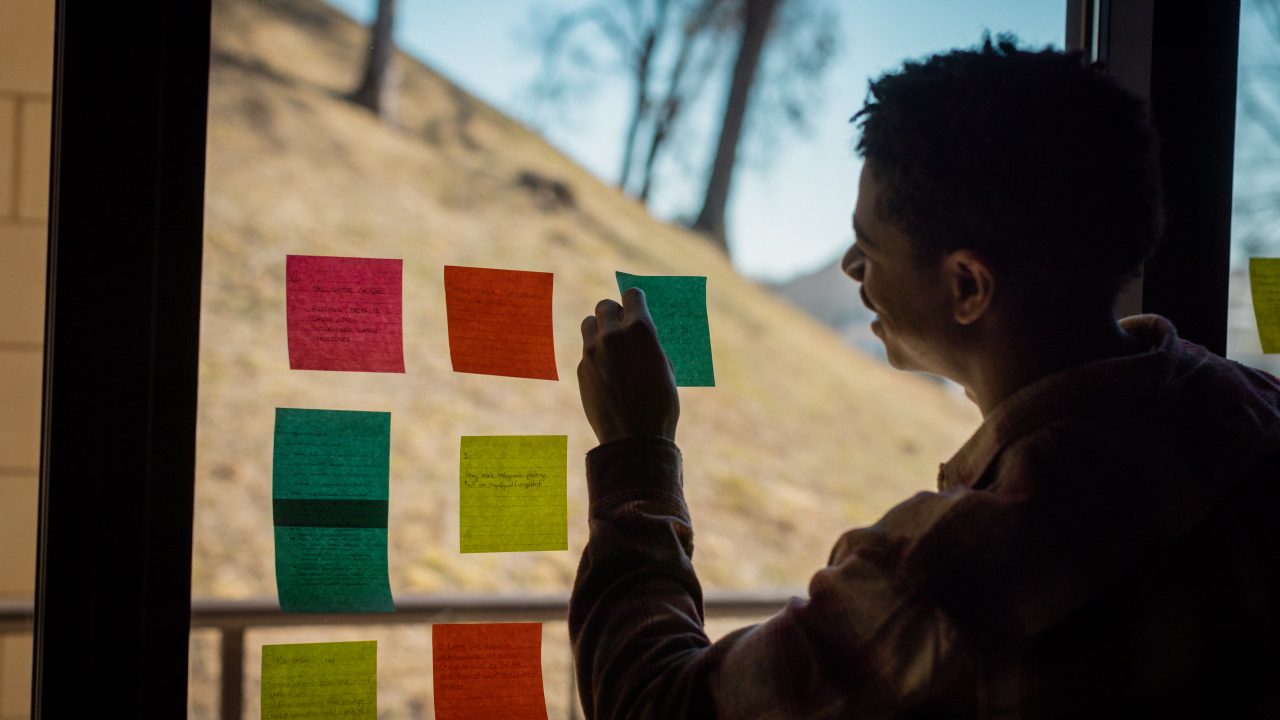 A person places colorful sticky notes with handwritten text on a window, with a hillside visible outside in the background.