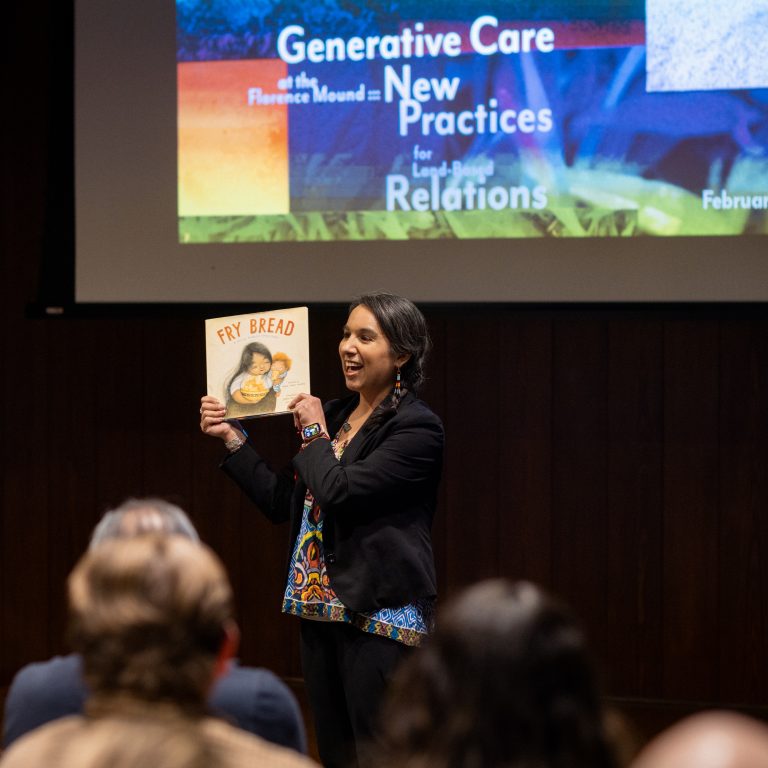 A woman stands in front of an audience holding up the book Fry Bread, with a presentation slide titled Generative Care: New Practices for Land-kin Relations displayed behind her.