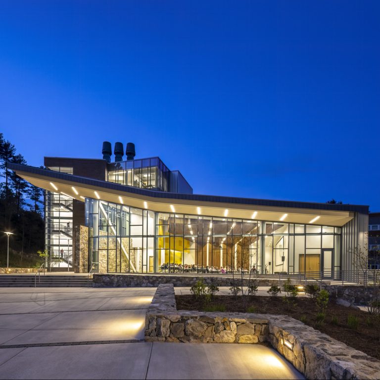 Modern glass and steel building illuminated at dusk, with a person walking in the foreground and landscaped stone walls bordering the walkway.
