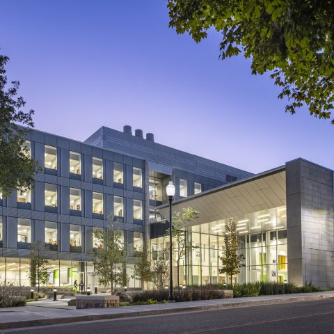 Modern four-story academic building with large glass windows, illuminated interior, and landscaped surroundings at dusk; trees and street in foreground.