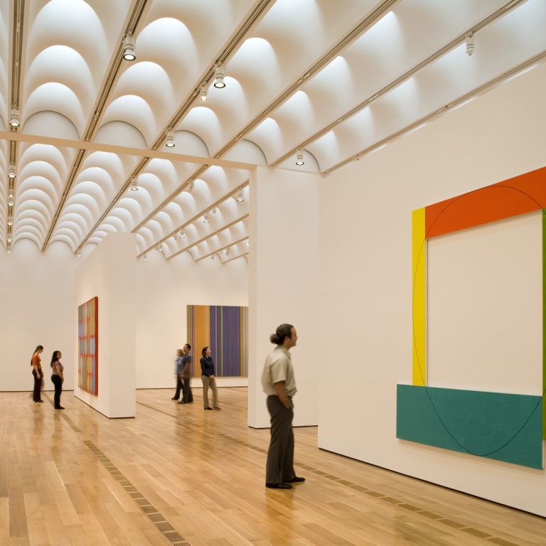 People observe colorful abstract paintings in a modern, well-lit art gallery with a wooden floor and a distinctive curved ceiling design.