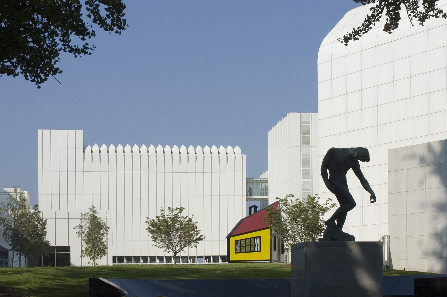 A modern art museum exterior with white geometric buildings, a yellow house sculpture, trees, and a bronze statue in the foreground.