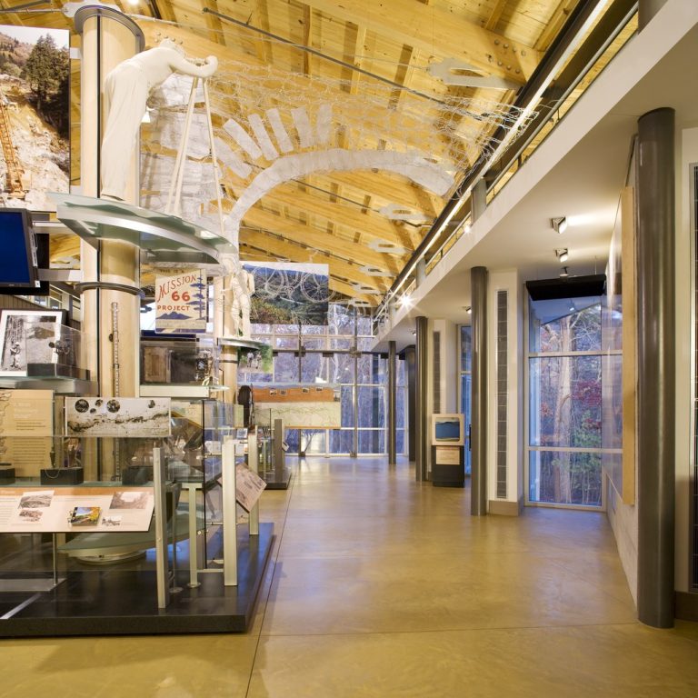 Museum interior with wooden ceiling, glass walls, and various exhibits including a white statue, informational panels, and Route 66 memorabilia displayed along a well-lit hallway.