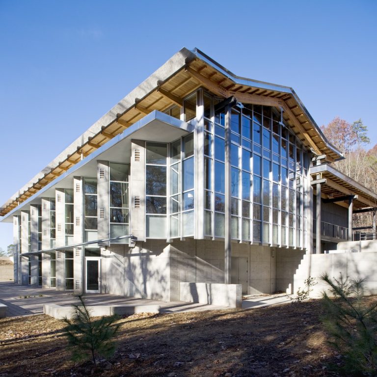 Modern building with large glass windows, exposed concrete, and a sloped wooden roof, set against a clear sky and surrounded by sparse trees.