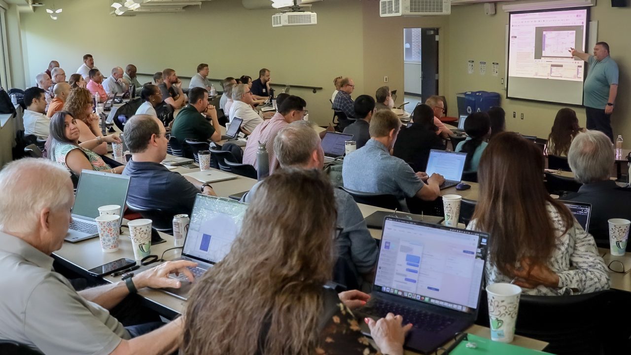 A group of people seated in a classroom listen to a presenter at the front, who points at a projected screen. Most attendees have laptops open.
