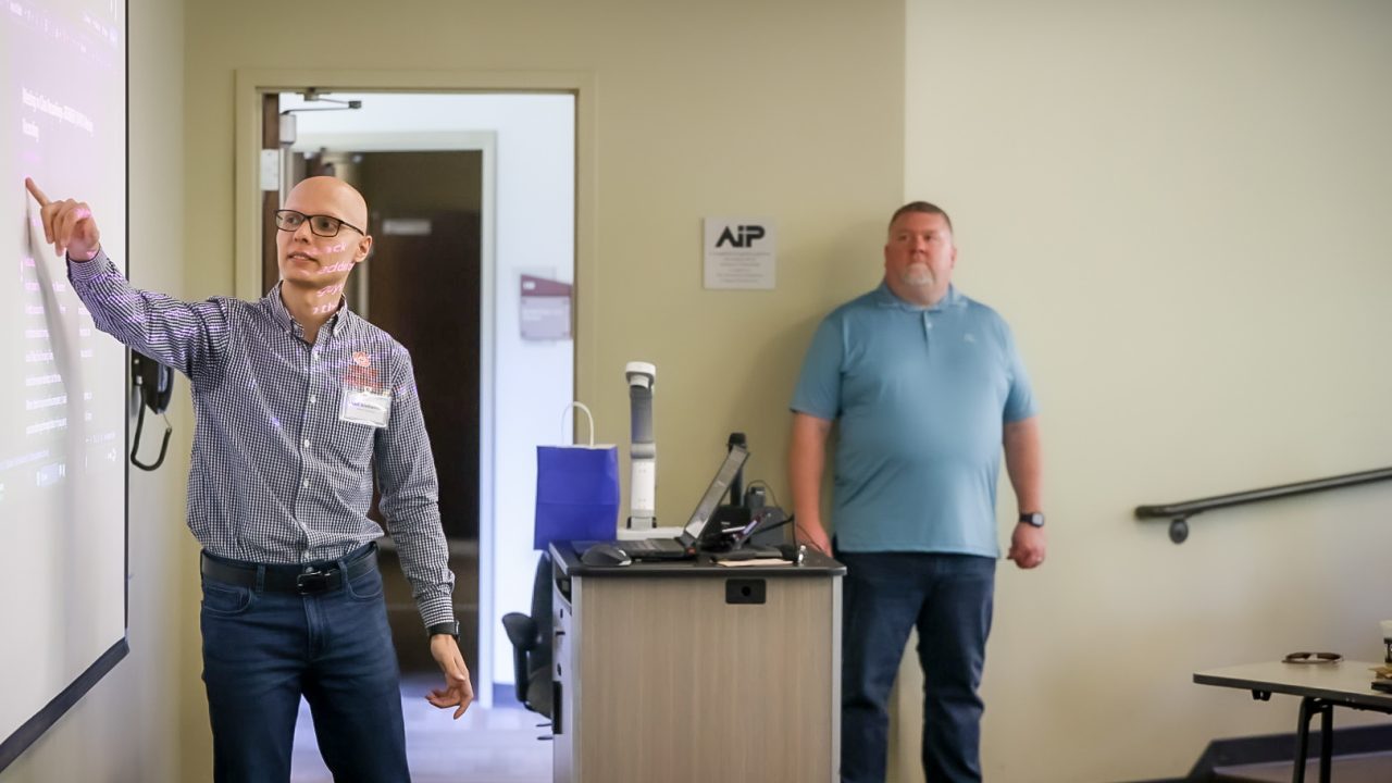 A man points at a projection screen during a presentation while another man stands by a lectern in a classroom setting.
