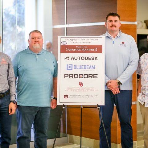 Four men stand beside a sign displaying sponsors for a construction symposium, including Autodesk, Bluebeam, and Procore, inside a building with glass walls.