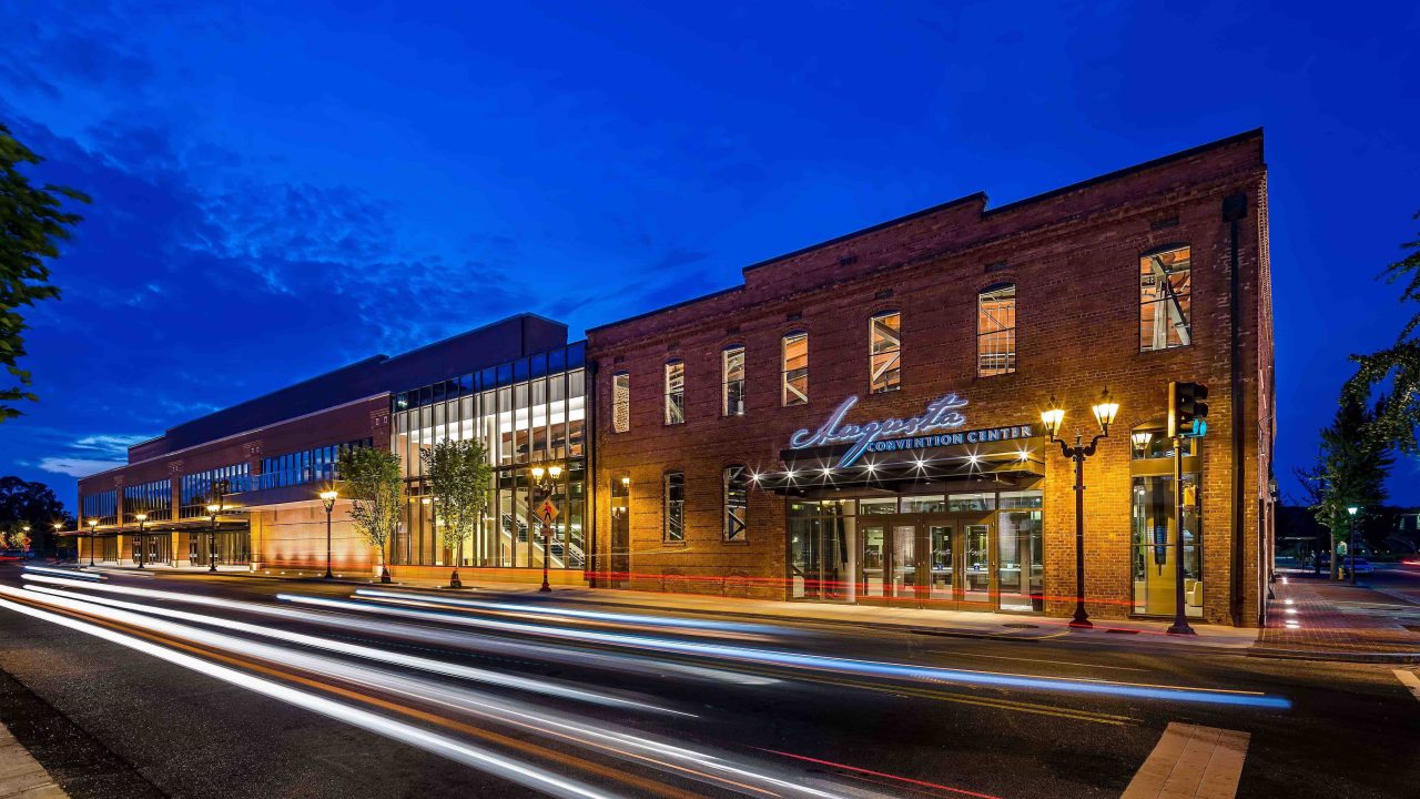 Exterior view of a brick convention center at dusk with illuminated signage and light trails from passing vehicles on the street.