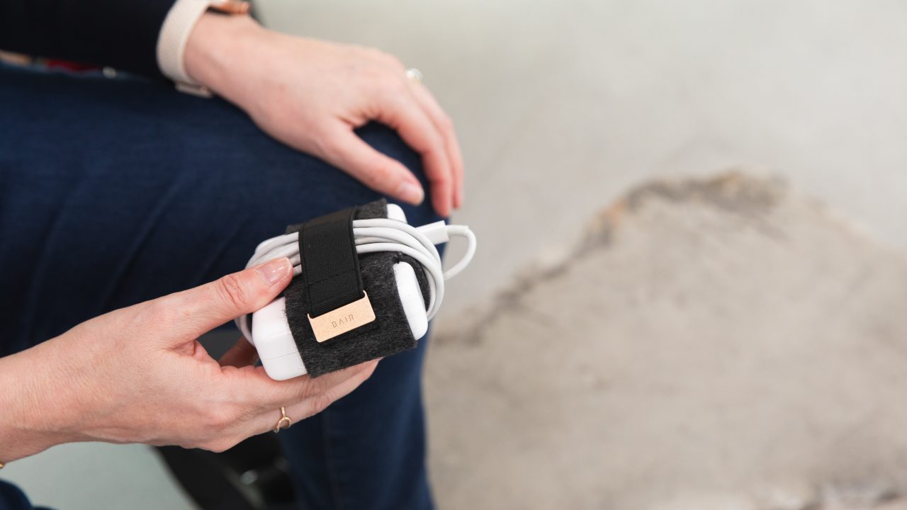 A person holds a neatly wrapped white charging cable secured with a black and brown strap while sitting on a light-colored surface.