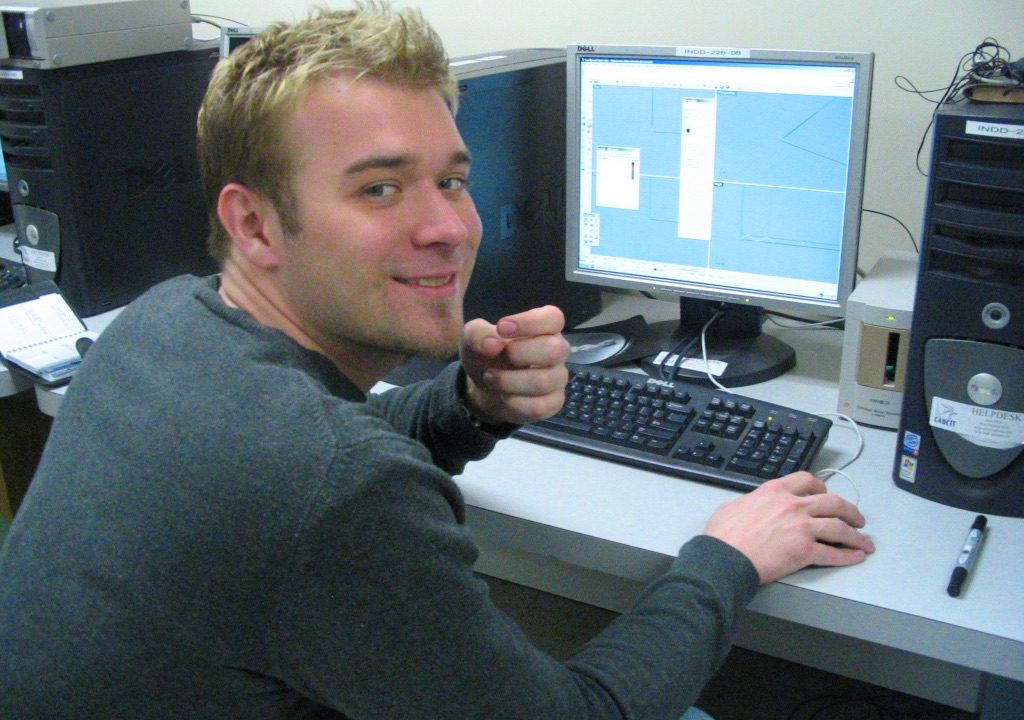 A person sits at a desk using a computer with design software open, turning to look at the camera and smiling. Multiple desktop towers and a keyboard are visible.