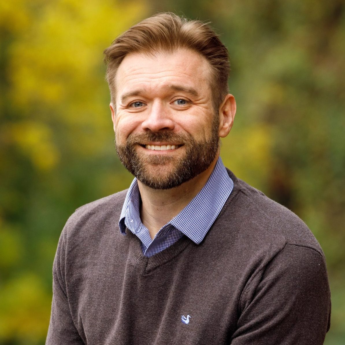 A man with a beard, wearing a brown sweater and blue shirt, sits outdoors on a stone surface with blurred greenery and autumn foliage in the background.