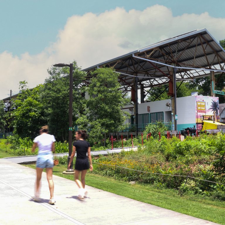 Three people walk on a concrete path near a landscaped area with tall green plants and a large, open-roofed structure in the background.