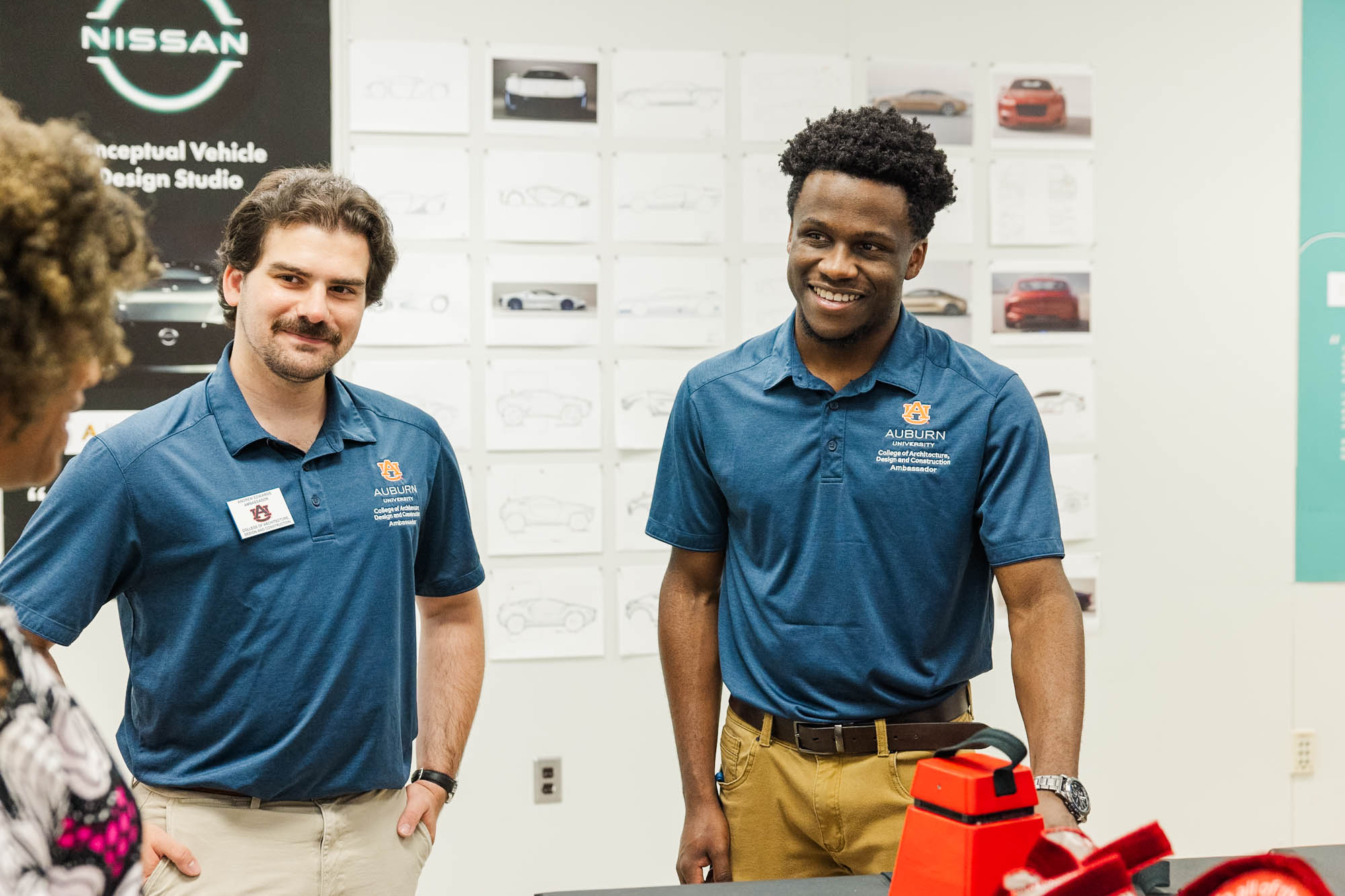 Two male students in blue Auburn University shirts stand indoors, smiling, with vehicle sketches and a red model visible in the background.