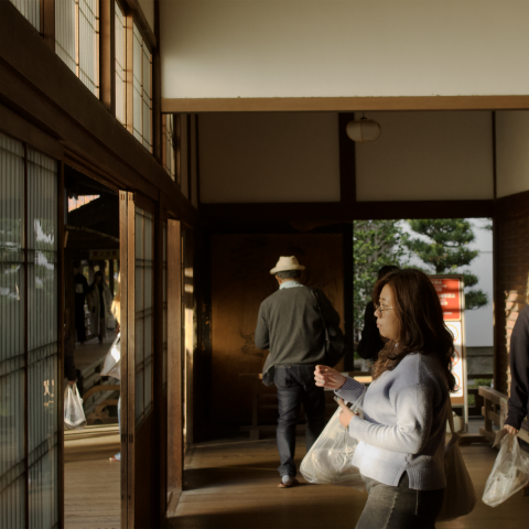 Three people walk through a hallway in a traditional Japanese building, carrying shopping bags. Sunlight streams through wooden-framed windows, casting shadows on the floor and walls.