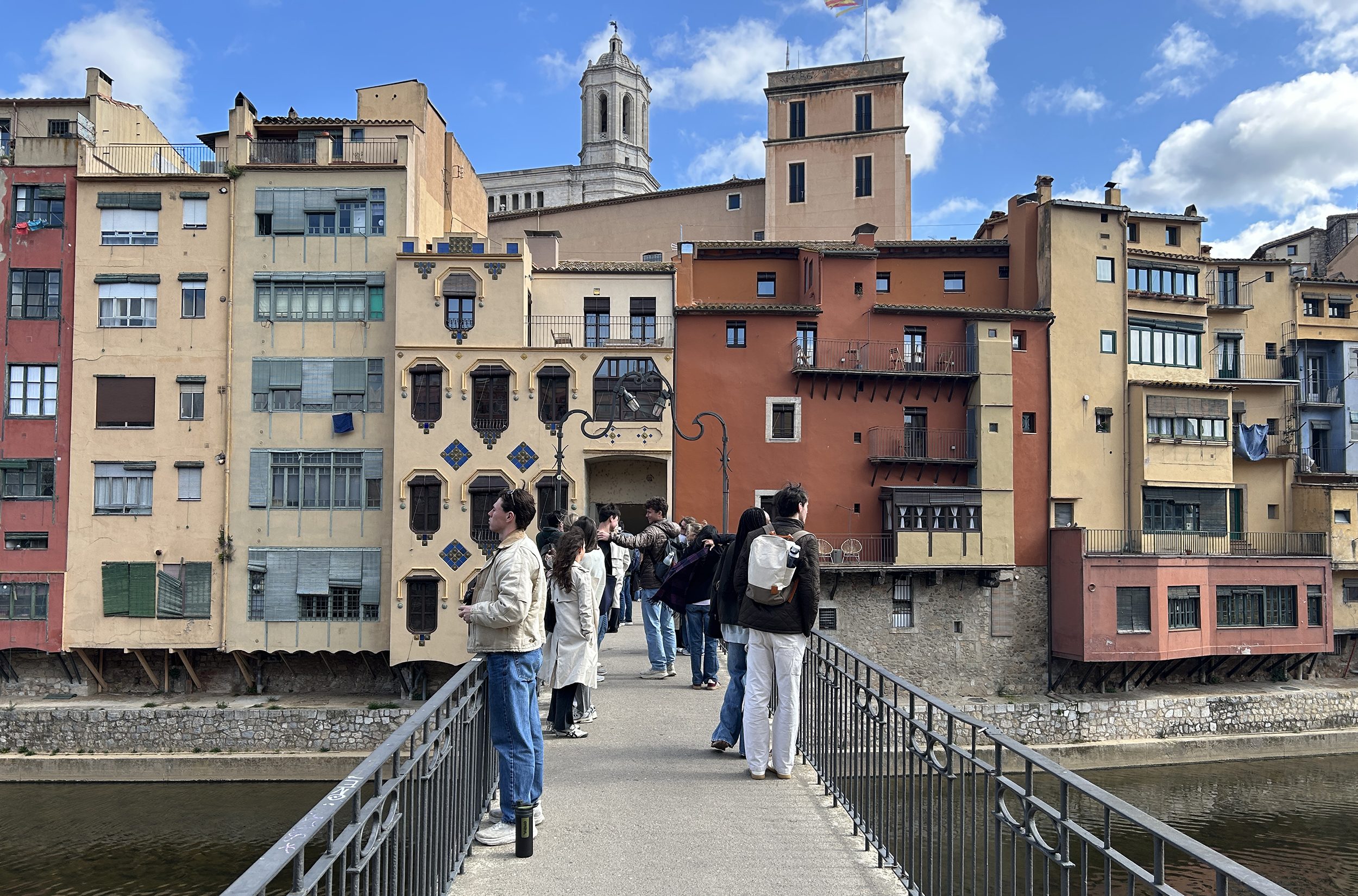 People stand and walk on a narrow pedestrian bridge over a river, with colorful buildings and a church tower in the background under a partly cloudy sky.
