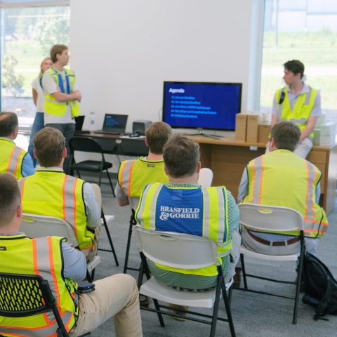 A group of people in yellow safety vests sit and listen to a presentation in a conference room, with a large agenda displayed on a monitor at the front.
