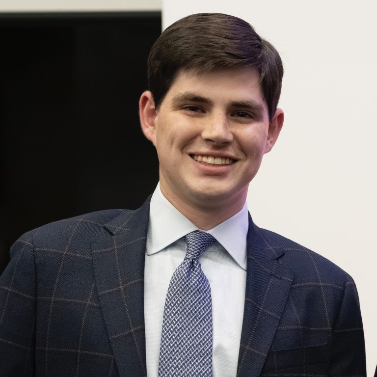 A man in a blue checked suit, light blue shirt, and patterned tie is smiling at the camera against a white and black background.