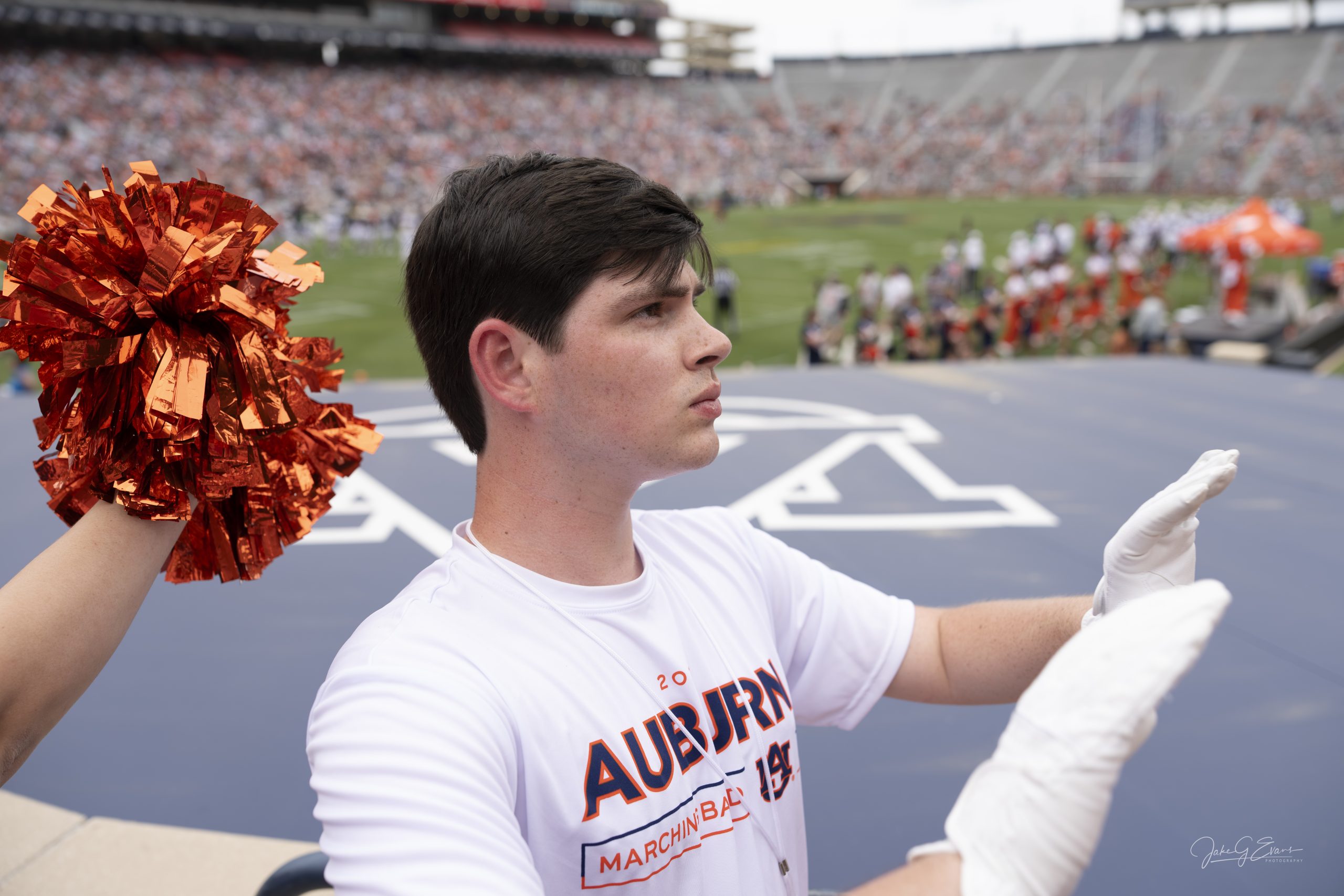 A person wearing an Auburn Marching Band shirt holds up gloved hands, with orange pom-poms and a football field visible in the background.