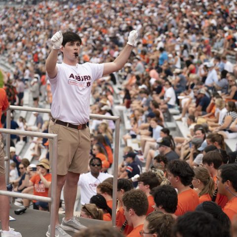 A person stands on a platform leading cheers at a crowded stadium, surrounded by people wearing orange and white shirts.