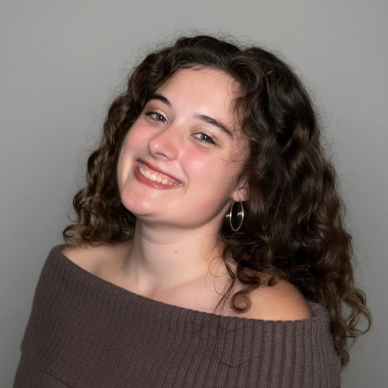 Young woman with curly hair wearing a brown off-the-shoulder sweater, smiling in front of a plain gray background.