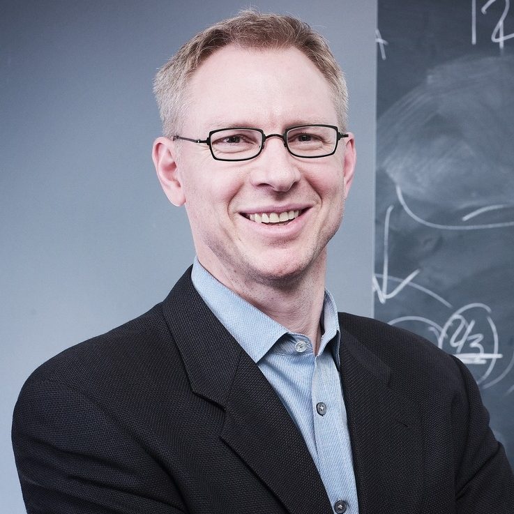 A man in a suit stands smiling with arms crossed in front of a blackboard covered with diagrams and sketches.