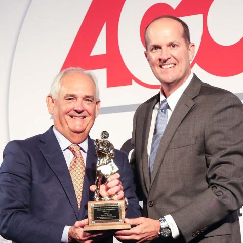 Two men in suits stand smiling; one hands an award trophy to the other in front of a backdrop with large red AGC letters.