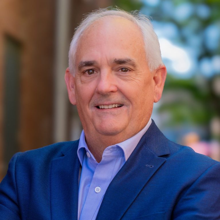 Older man with white hair in a blue suit stands outdoors with arms crossed, smiling at the camera; blurred brick wall and greenery in background.
