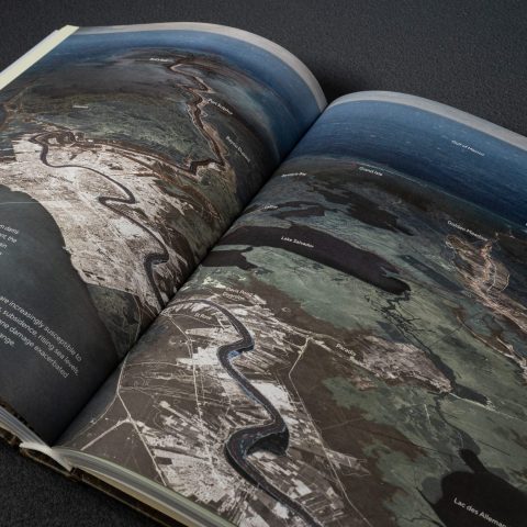 An open book displays an aerial map with labeled features, including rivers, lakes, and coastline, alongside descriptive text on the left page.